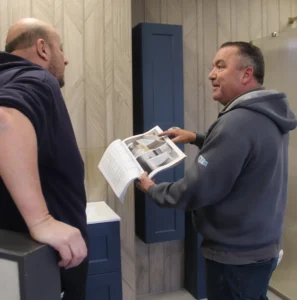 Two men stood talking to each other in a showroom. The man on the right is holding a catalogue open and pointing to an image of a bathroom.